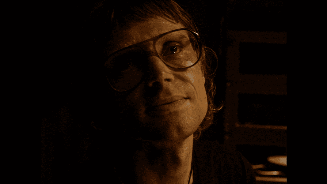 Who's Watching 4 Woman wearing oversized glasses in a dim, sepia-toned room, looking upward with a soft smile under warm lighting.