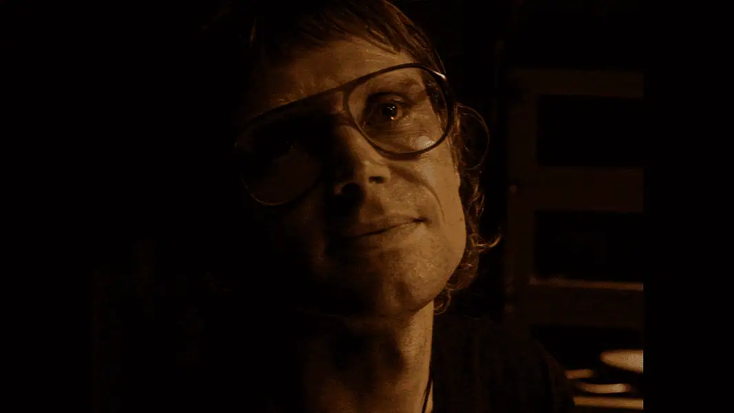 Who's Watching 4 Woman wearing oversized glasses in a dim, sepia-toned room, looking upward with a soft smile under warm lighting.