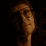OFX hosting hyper-modern horror pic from indie-rock hero Woman wearing oversized glasses in a dim, sepia-toned room, looking upward with a soft smile under warm lighting.