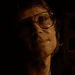 OFX hosting hyper-modern horror pic from indie-rock hero Woman wearing oversized glasses in a dim, sepia-toned room, looking upward with a soft smile under warm lighting.
