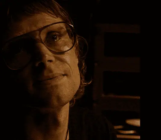 OFX hosting hyper-modern horror pic from indie-rock hero Woman wearing oversized glasses in a dim, sepia-toned room, looking upward with a soft smile under warm lighting.