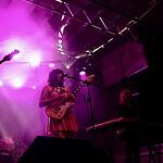 Band performing on a lit stage with purple lights; two guitarists and a keyboard player, drum kit in back, vintage TV on a red pedestal to the left.