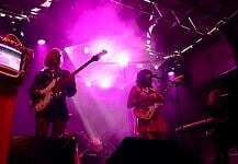 Catch these scene-leading locals opening for can’t-miss nationals across OKC Band performing on a lit stage with purple lights; two guitarists and a keyboard player, drum kit in back, vintage TV on a red pedestal to the left.