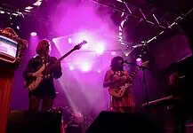 Catch these scene-leading locals opening for can’t-miss nationals across OKC Band performing on a lit stage with purple lights; two guitarists and a keyboard player, drum kit in back, vintage TV on a red pedestal to the left.