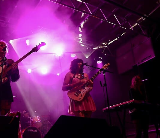 Catch these scene-leading locals opening for can’t-miss nationals across OKC Band performing on a lit stage with purple lights; two guitarists and a keyboard player, drum kit in back, vintage TV on a red pedestal to the left.